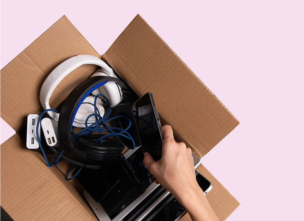 A light skinned man reaching into a cardboard box full of electrical goods against a pink background.
